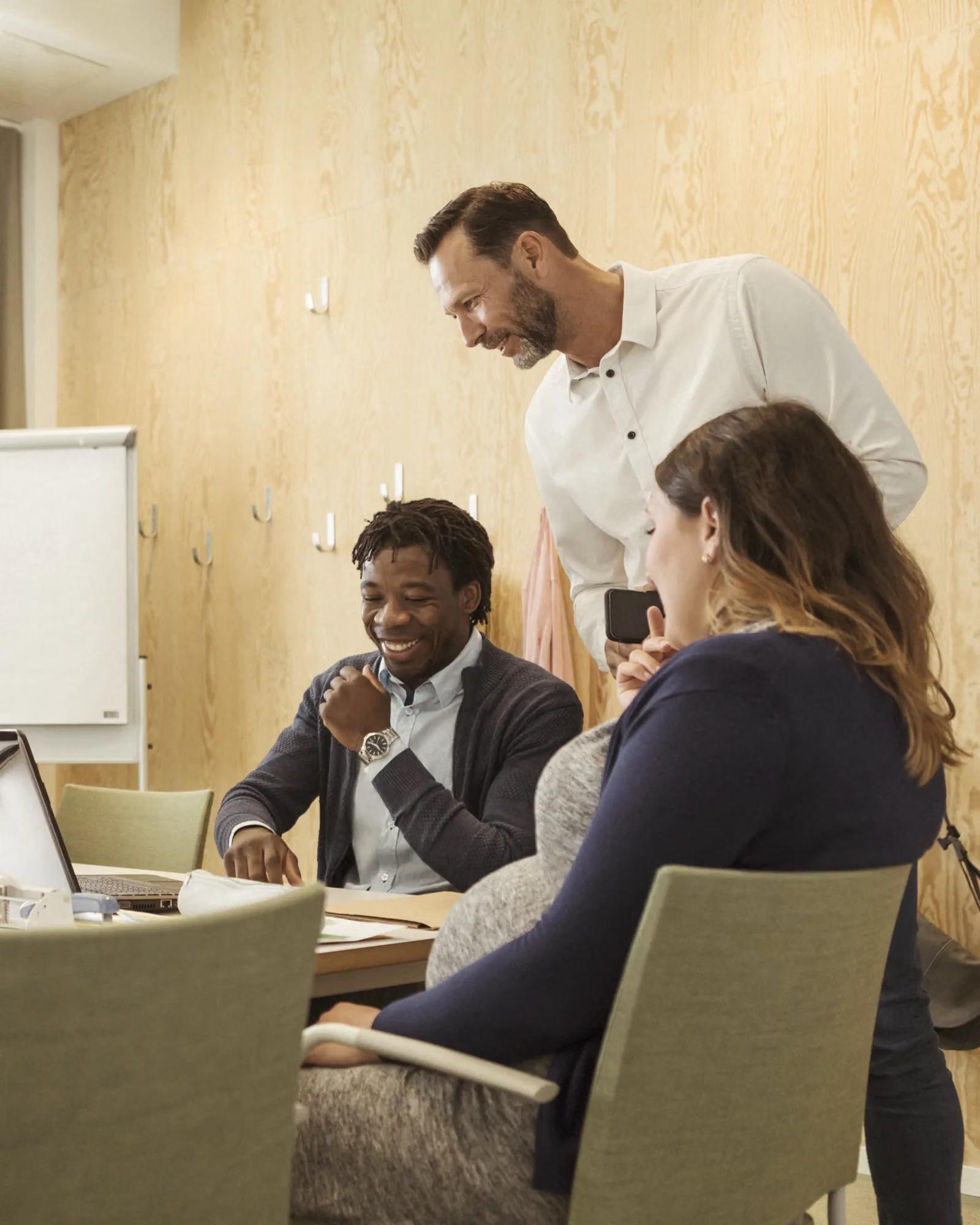 Three colleagues, including a pregnant woman, smile while collaborating around a laptop in a meeting.