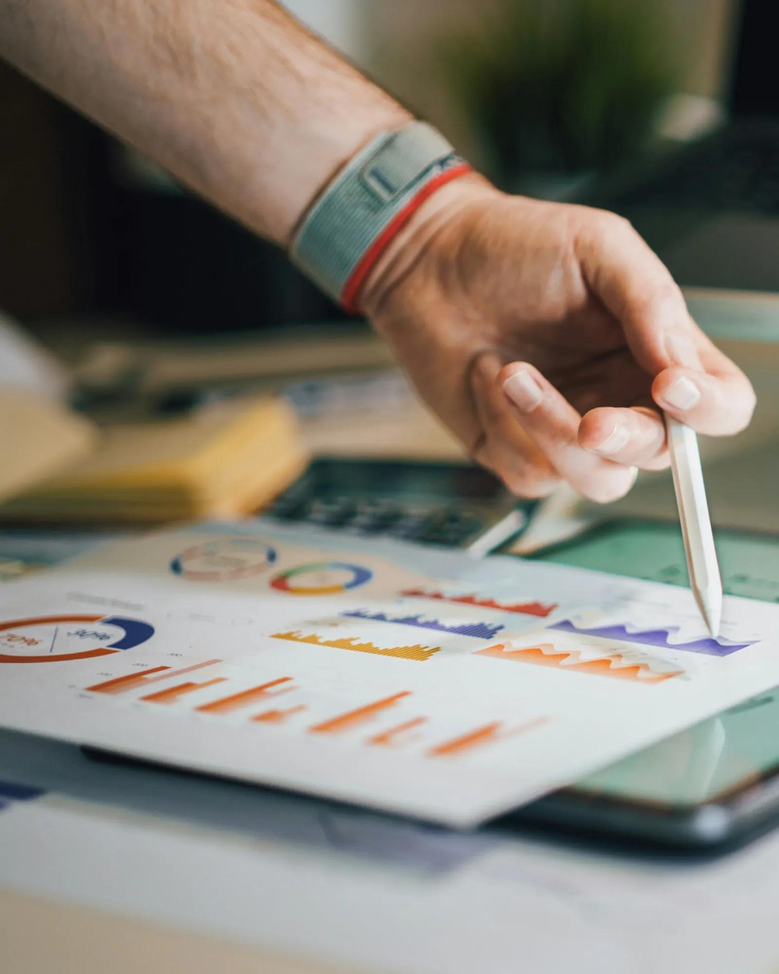 A hand uses a stylus to point at charts and graphs on a tablet, showing data analysis or performance reporting on a desk.