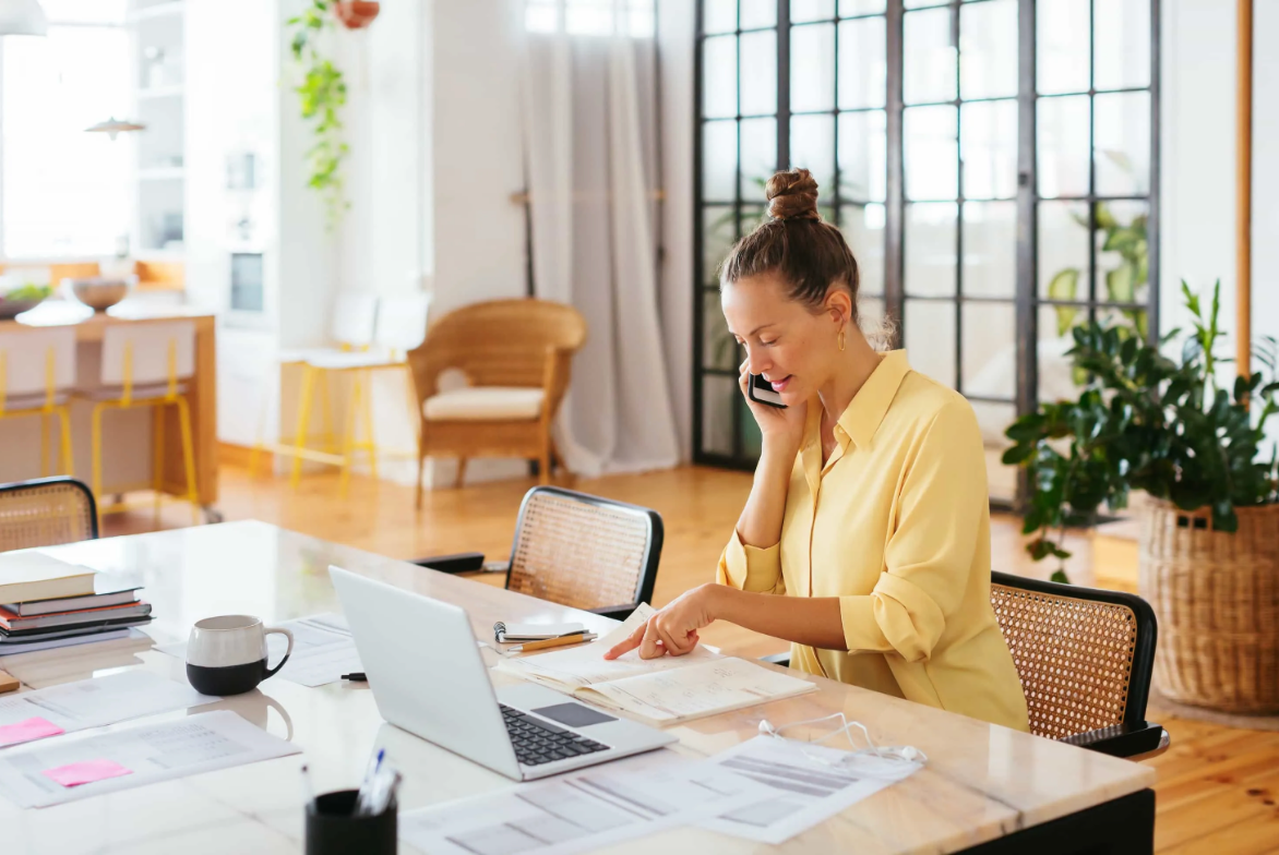 A woman talks on a phone while pointing at documents next to a laptop on a table.