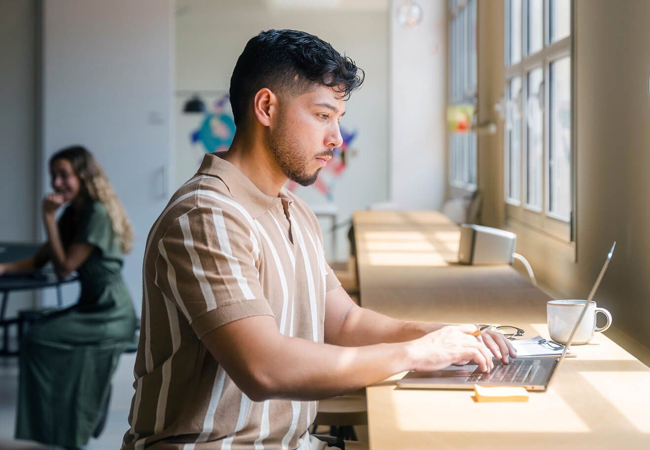 A man types on a laptop at a sunlit desk by a window.