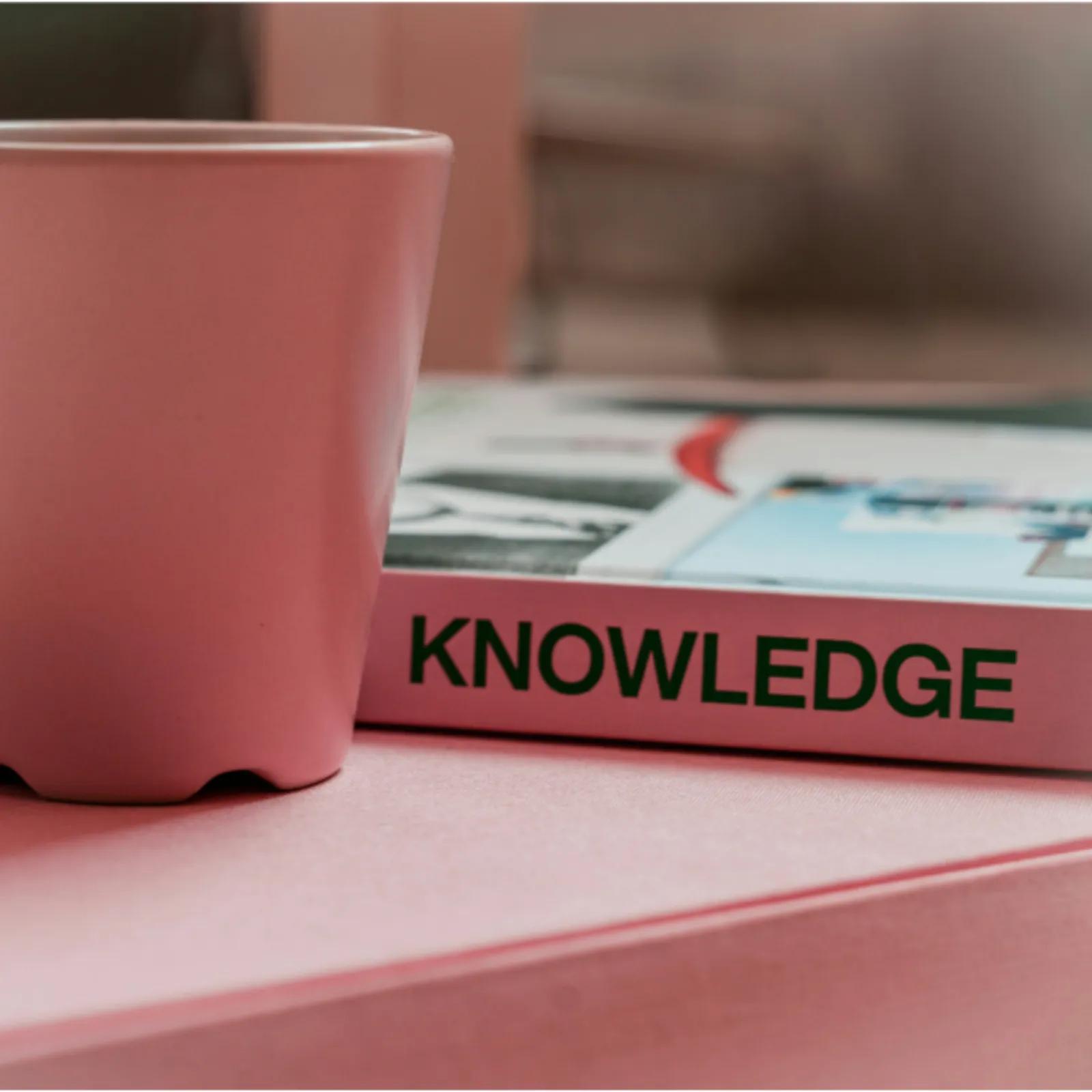 A pink coffee cup sits beside a book titled “Knowledge” on a desk