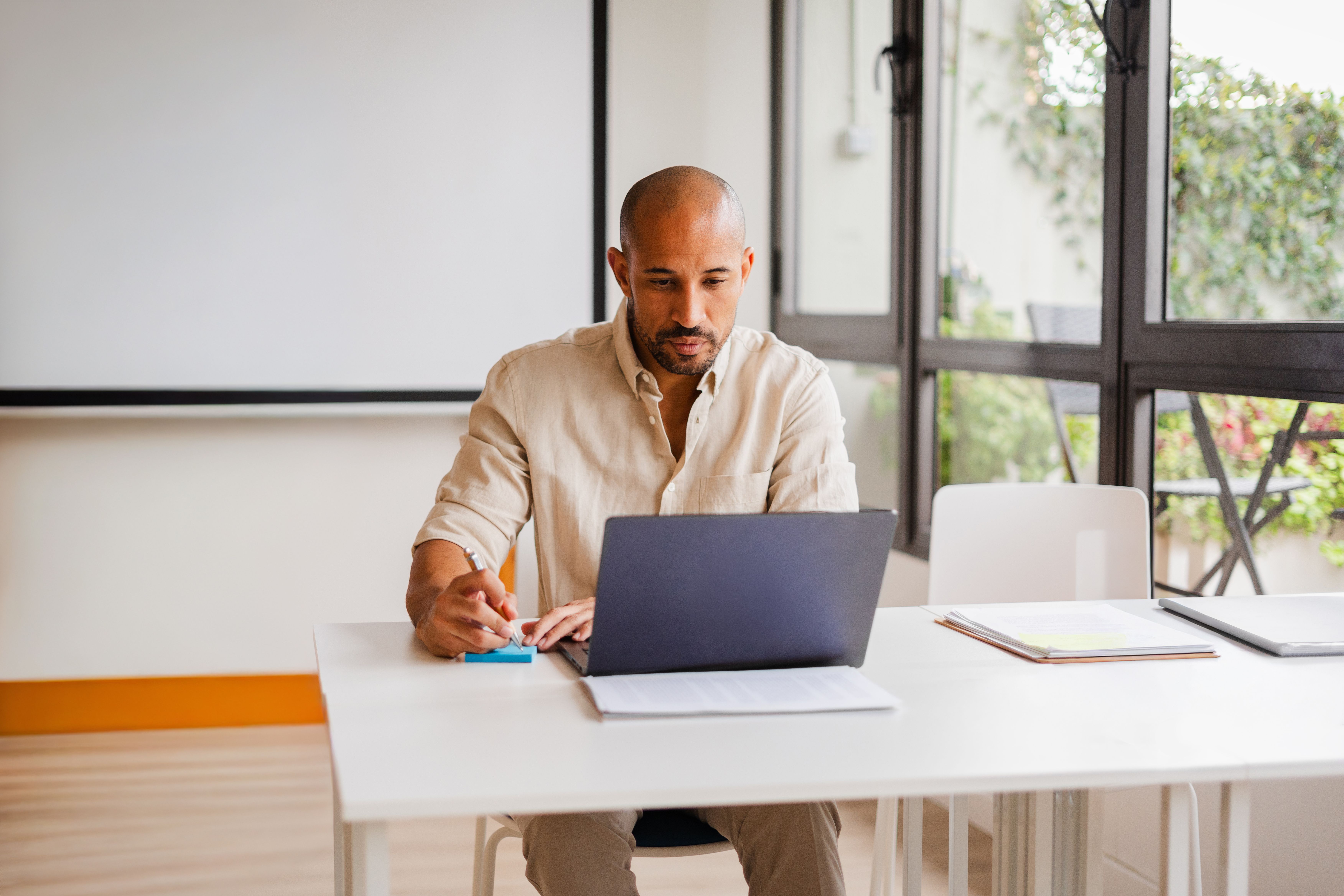 A bald man with a beard works on a laptop at a desk in a room with a projector screen and windows.