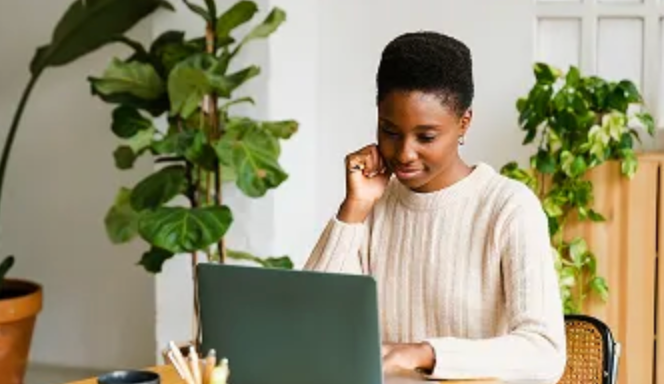 A young Black woman focused on a laptop at a desk, with plants in the background.