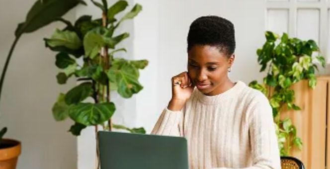 A young Black woman focused on a laptop at a desk, with plants in the background.