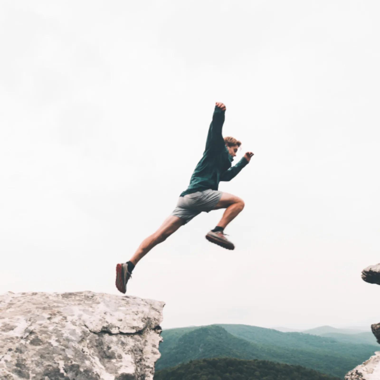 A person jumps between two rocky ledges high above a mountain landscape