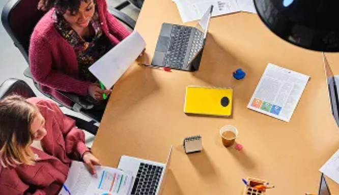 Overhead view of two women working together at a desk with laptops and documents.