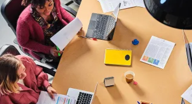 Overhead view of two women working at a table with laptops and documents.