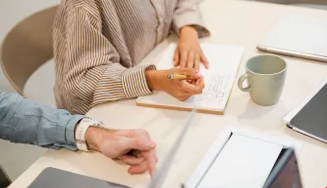 Two people working at a table, one writing in a notebook.