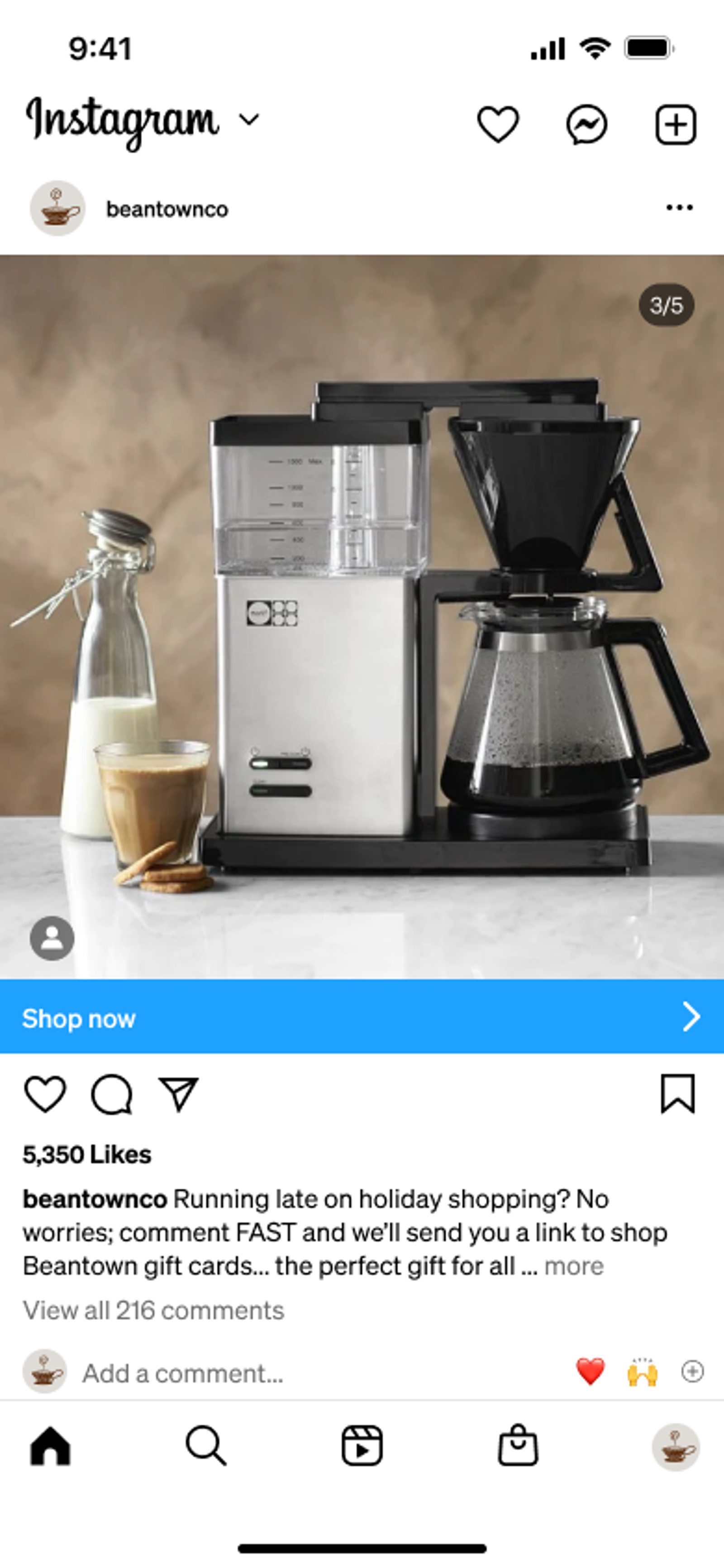 A silver and black coffee maker with a carafe of coffee, next to a glass of latte, a bottle of milk, and cookies on a marble countertop.