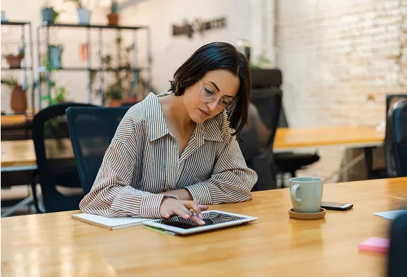 A woman with glasses uses a tablet at a wooden desk.