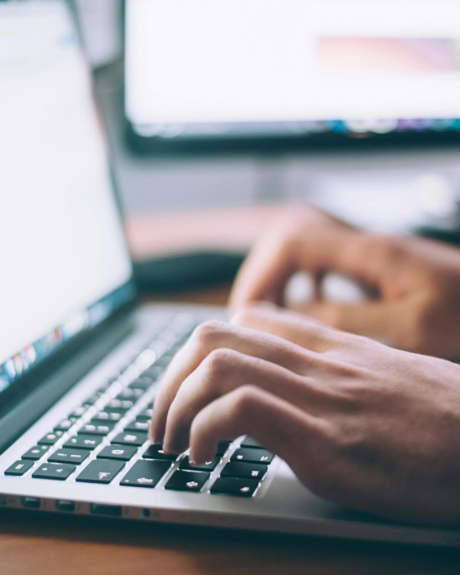 Hands type on a laptop keyboard at a desk, with a blurred computer screen in the background.