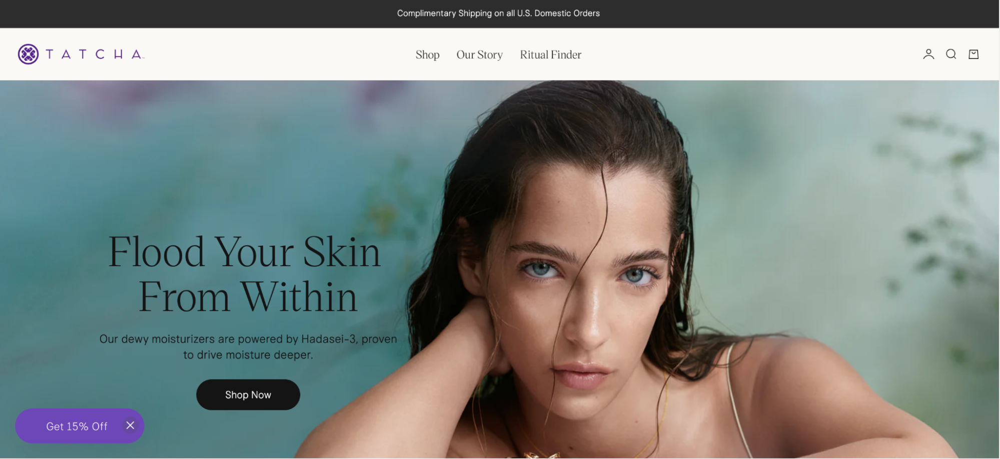 Image shows a screenshot of the Tatcha homepage, which features a hero image of a model with wet hair gazing into the camera against a backdrop of blues, purples, and greens. The copy across the image reads, “Flood your skin from within: our dewy moisturizers are powered by Hadasei-3, proven to drive moisture deeper.” The copy is followed by a “Shop now” CTA button.