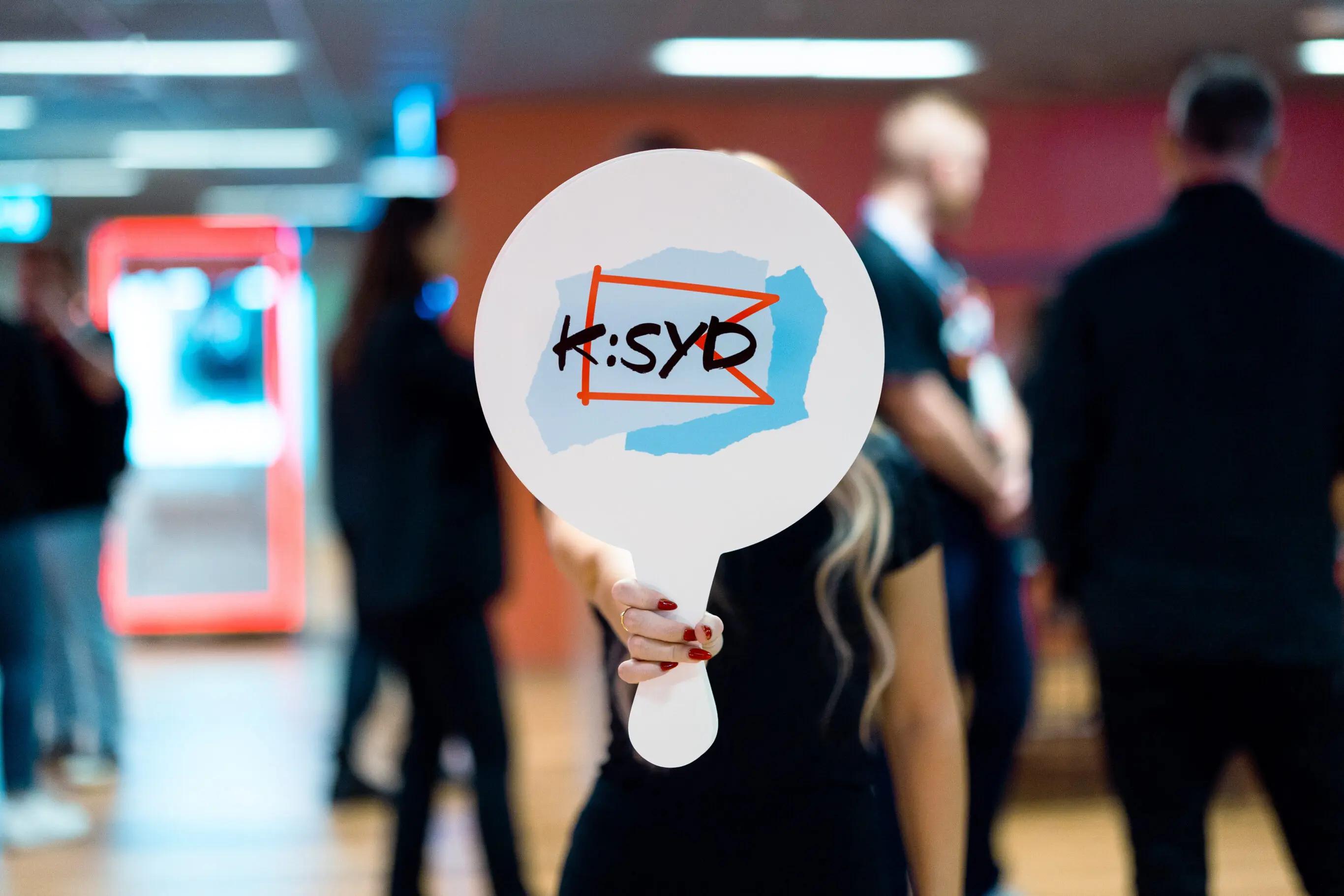 Person holding a sign with "K:SYD" written on it, in a blurred indoor setting with people in the background.