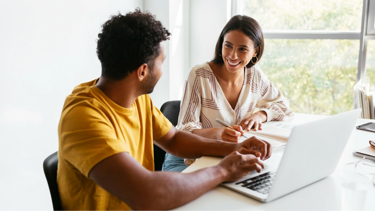 Two coworkers smile and collaborate at a desk with a laptop and notebook.