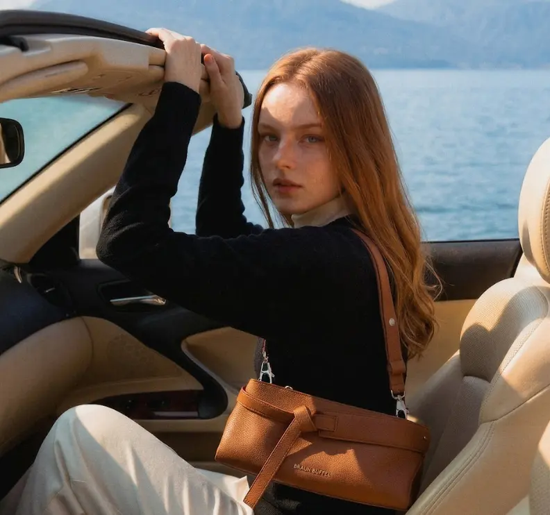 A red-haired woman in a black top and light pants sits in a convertible with a brown leather crossbody bag, looking at the viewer, with water and mountains in the background.