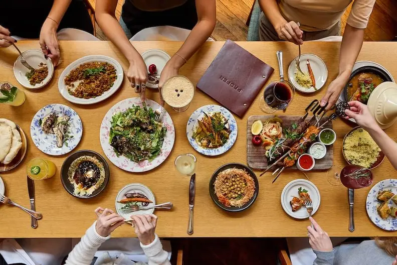 Overhead view of several people sharing a large spread of Middle Eastern food and drinks on a wooden table.
