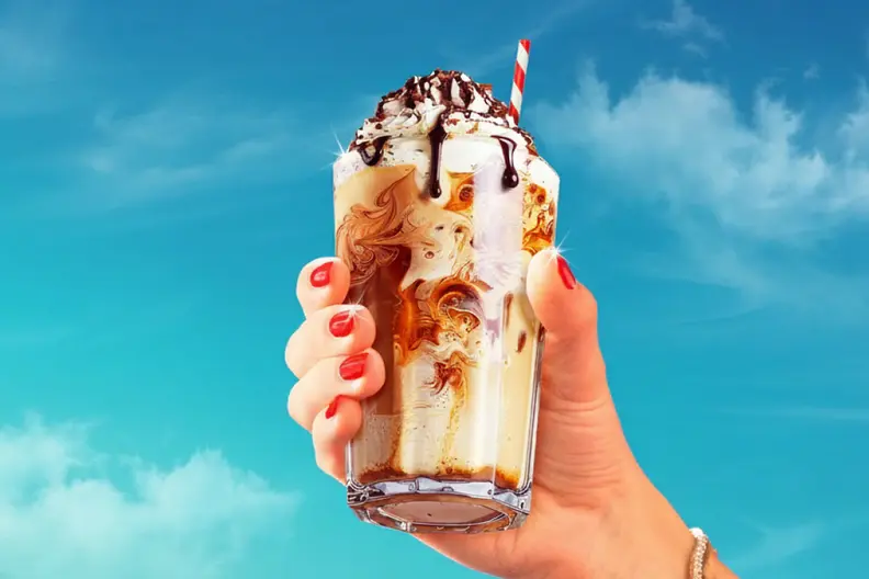 A hand with red nails holds an iced coffee topped with whipped cream, chocolate drizzle, and a straw, against a blue sky.