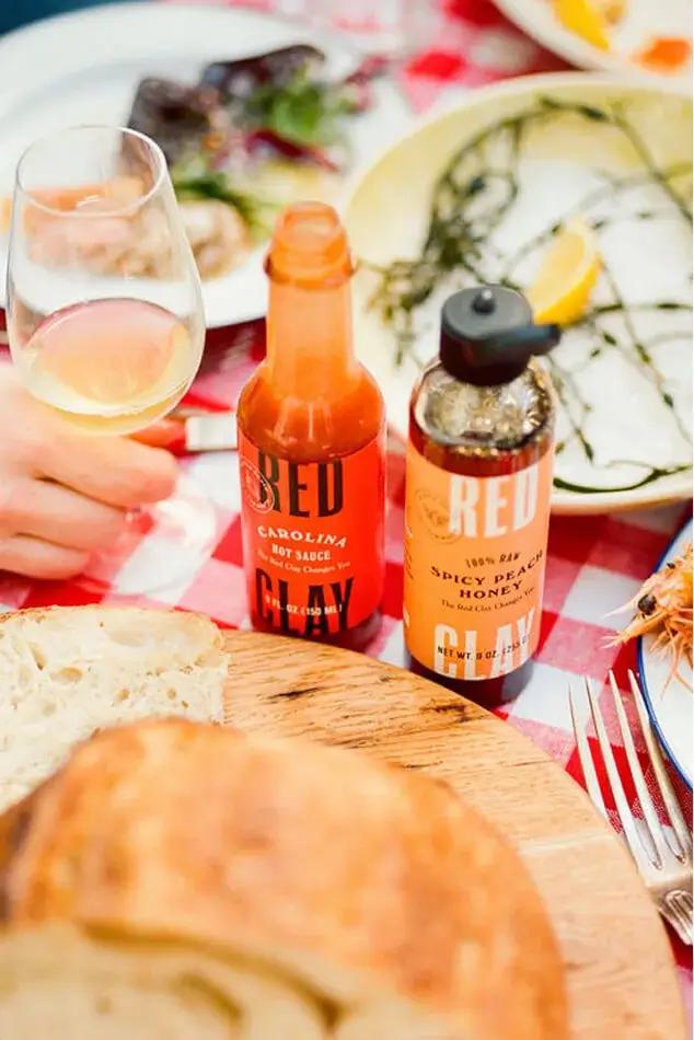 Bottles of Carolina hot sauce and spicy peach honey on a picnic table with bread, plates and a glass of wine.