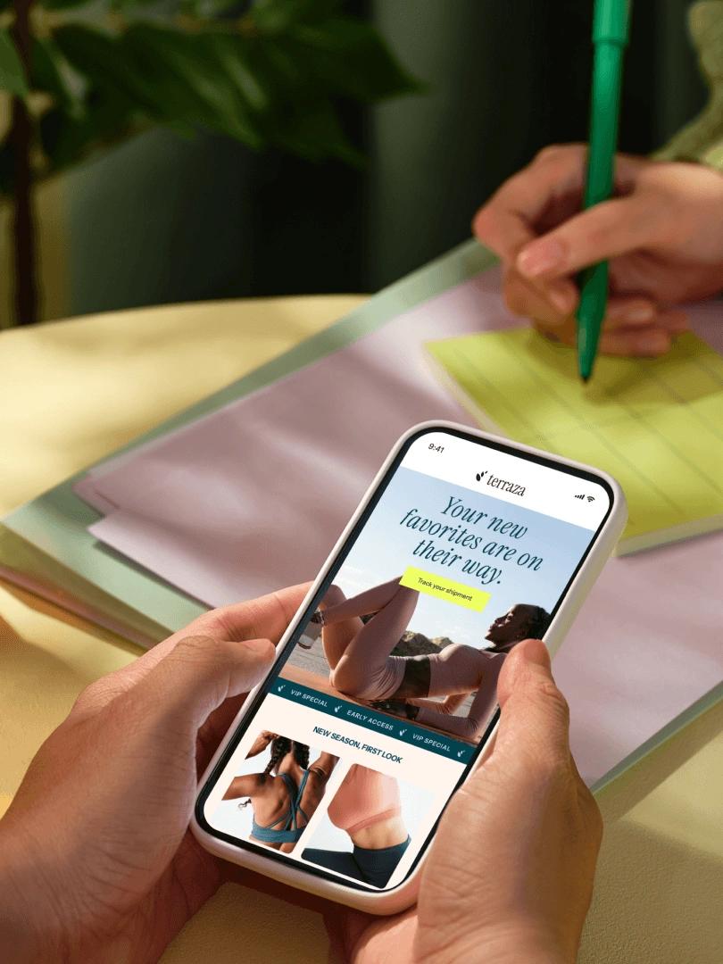 Person browses activewear on a smartphone while another writes on a notepad in the background.