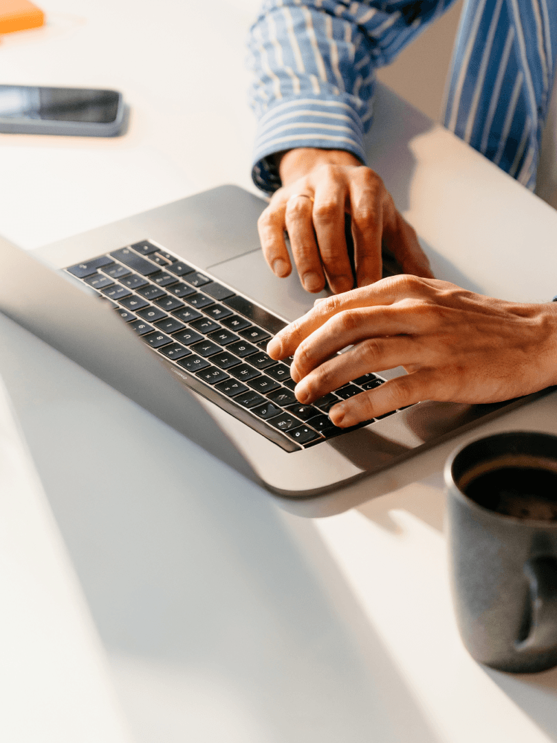 Person in a striped shirt typing on a laptop beside a mug of coffee.