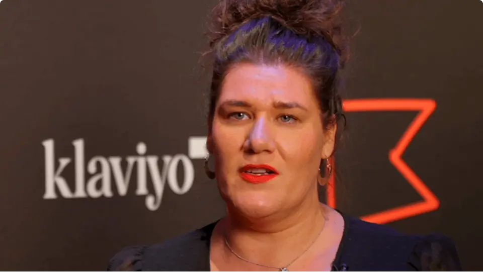 A woman with red lipstick speaks in front of a backdrop featuring the Klaviyo logo.