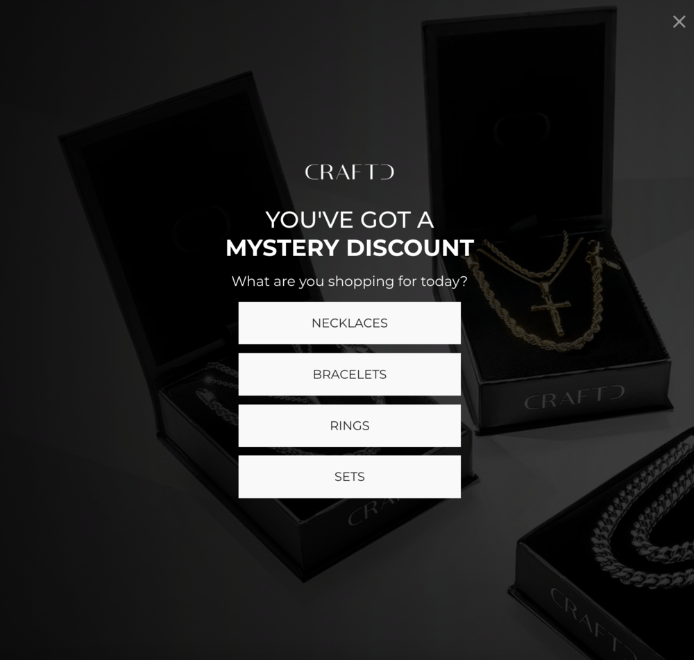 Modal popup from CRAFTD jewelry brand with a headline that says, “You've got a mystery discount”. Underneath is a question, “What are you shopping for today?” followed by 4 white buttons for product categories: necklaces, bracelets, rings, and sets. The background shows black luxury jewelry boxes with CRAFTD branding and chain jewelry pieces in dark, premium product photography style.