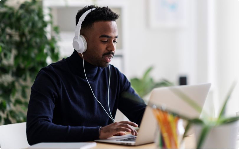 Man wearing headphones works on a laptop at a desk in a bright, plant-filled room.