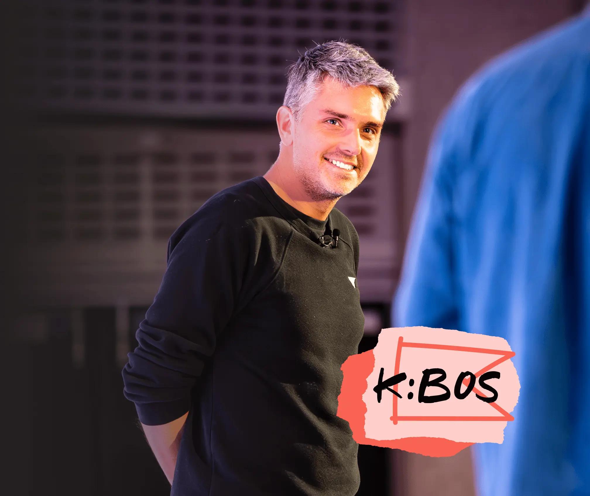 Smiling man in a black shirt with a microphone, standing indoors. Text overlay reads "K:BOS.