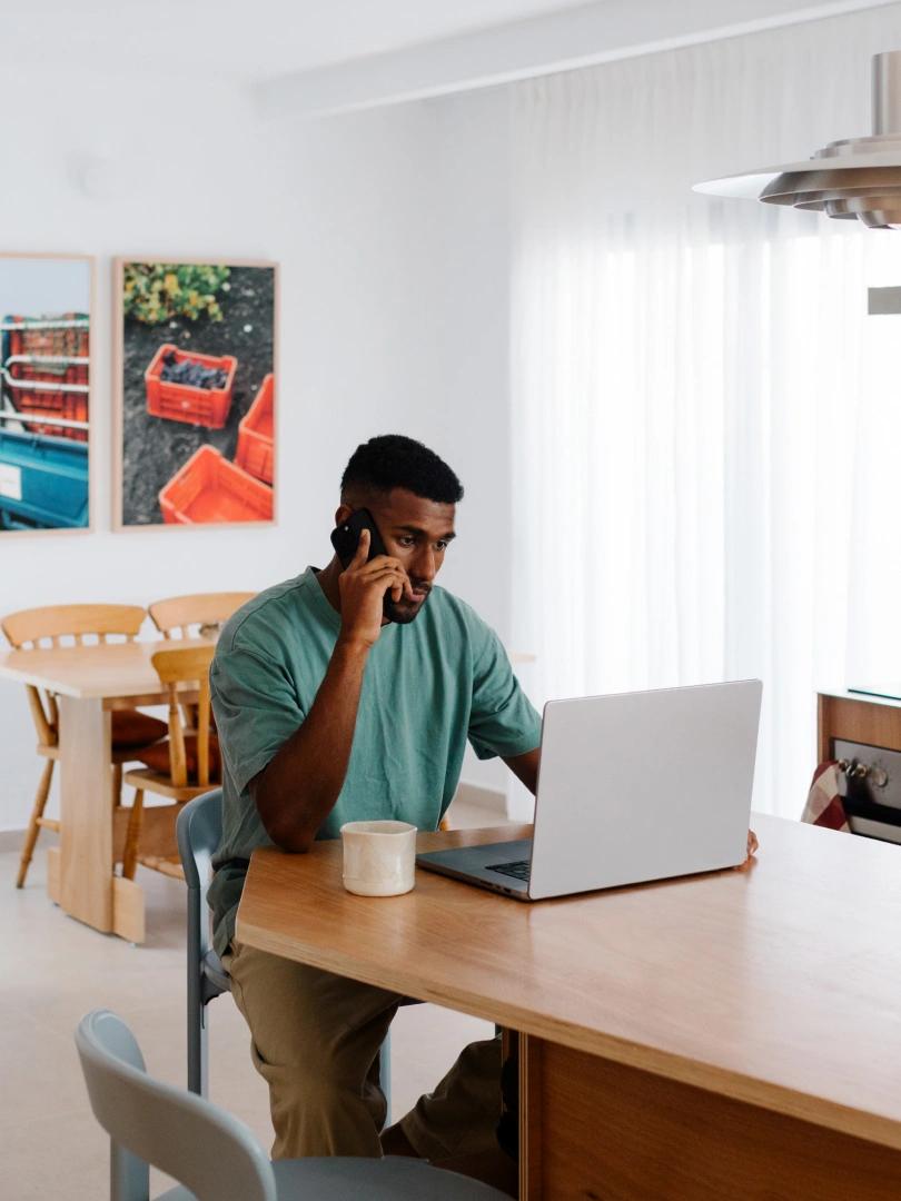 Man talks on the phone while working on a laptop at a kitchen table.
