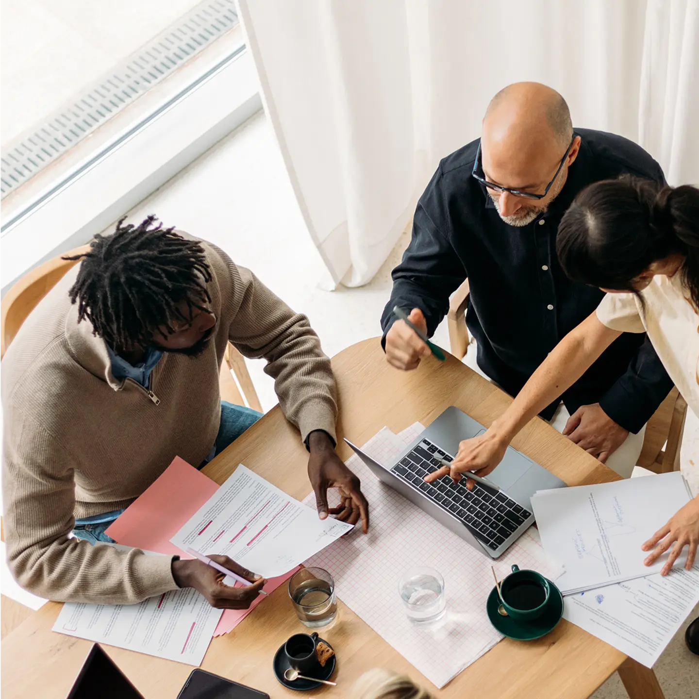Drei Personen arbeiten an einem Tisch mit Laptop, Dokumenten und Getränken. Eine Person zeigt auf den Laptopbildschirm.