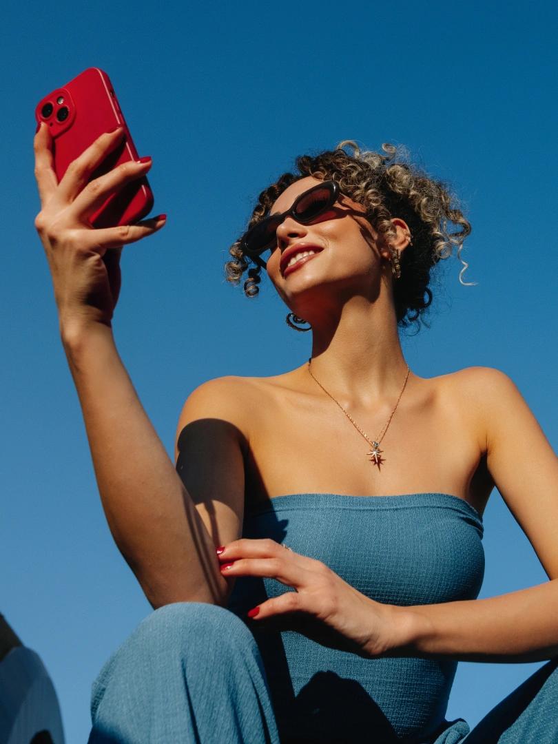 Smiling woman in sunglasses takes a selfie with a red phone against a clear blue sky.