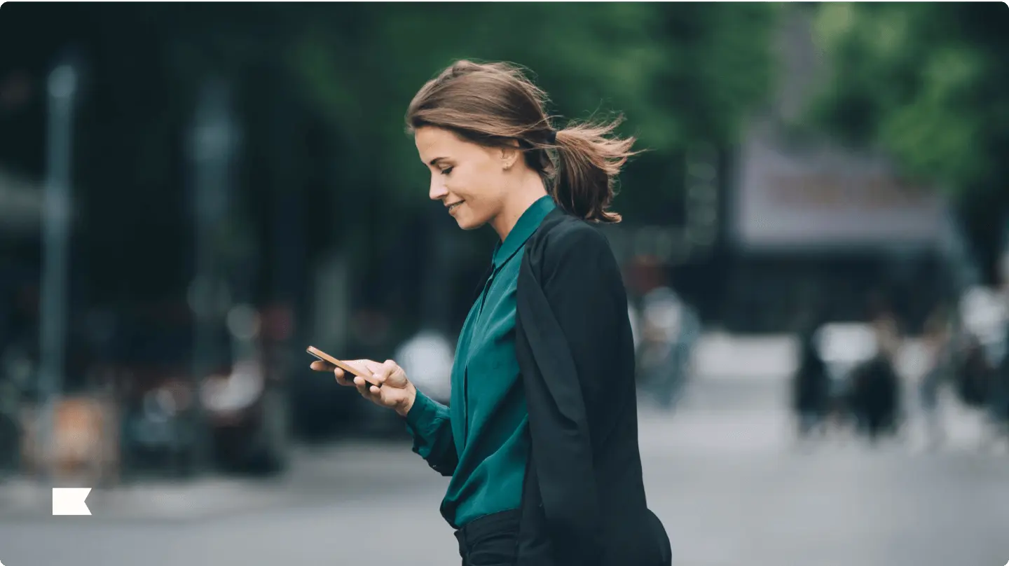 Woman walking outdoors, looking at her phone, with a blurred city background.