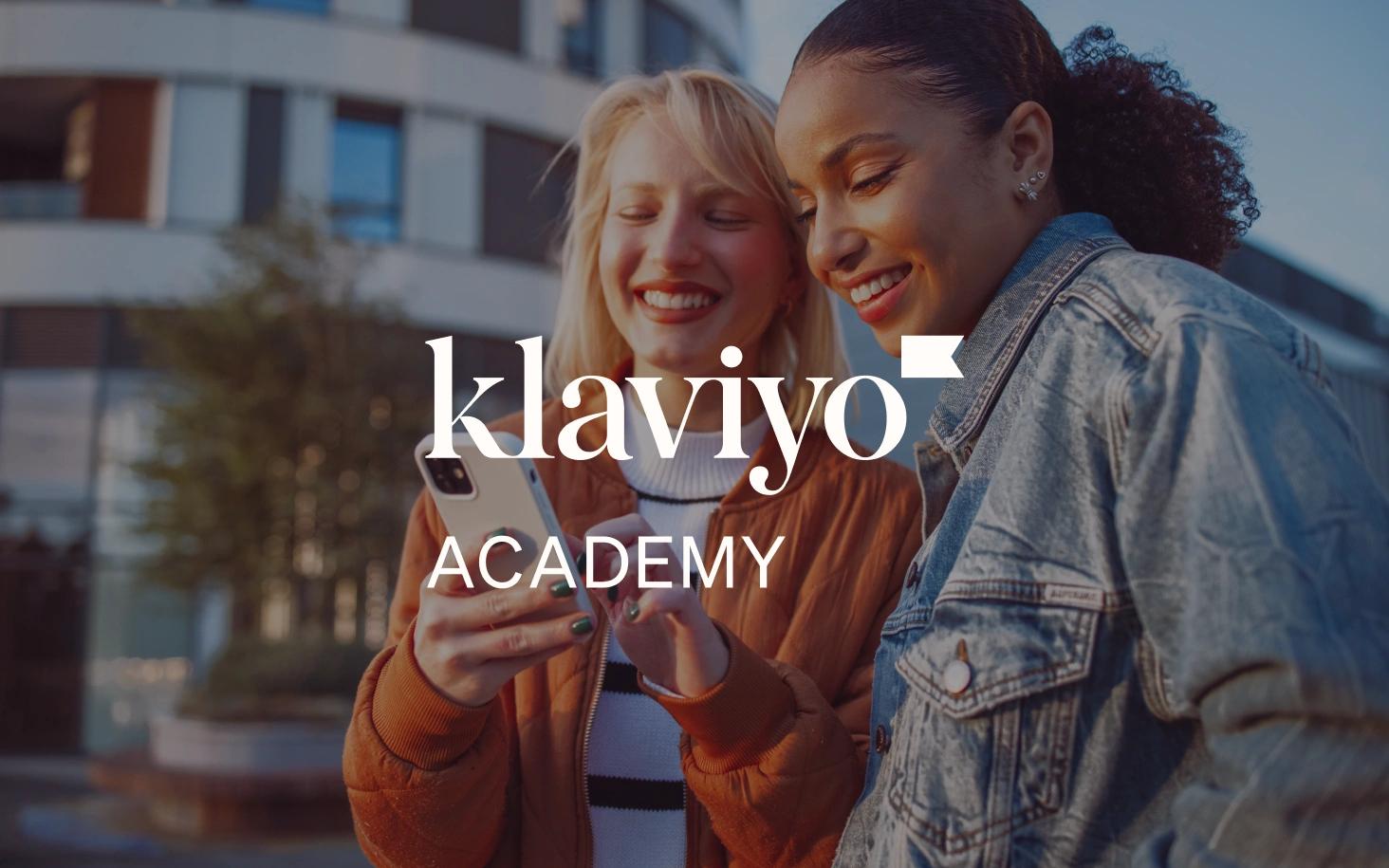Two smiling women look at a smartphone outdoors, overlaid with the Klaviyo Academy logo.