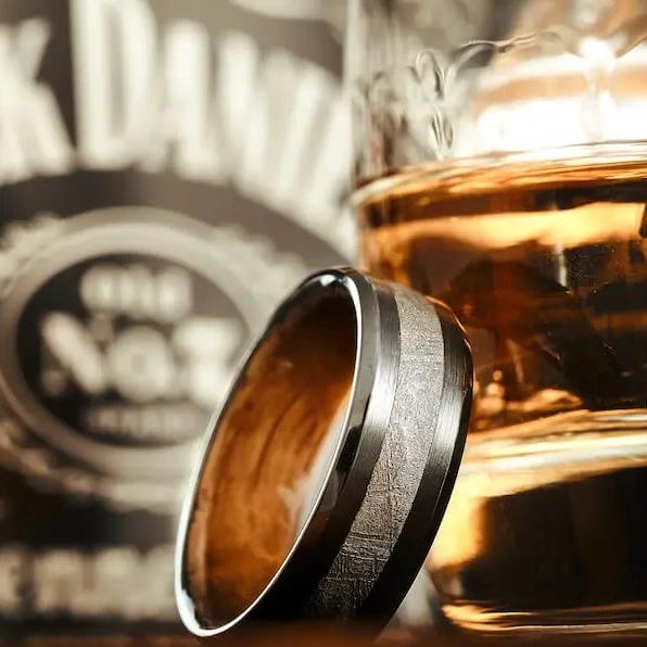 Black metal ring leaning against a glass of amber whiskey, with a blurred whiskey barrel in the background.