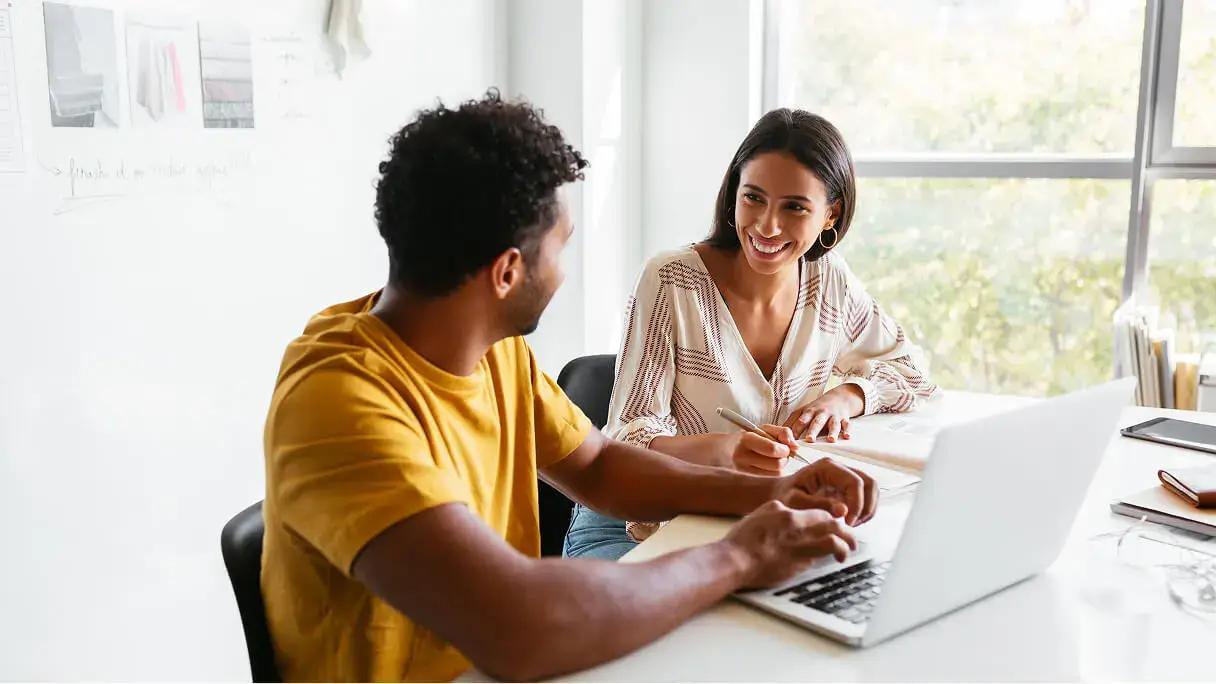 Two coworkers smile and collaborate at a desk with a laptop and notebook in a bright office.