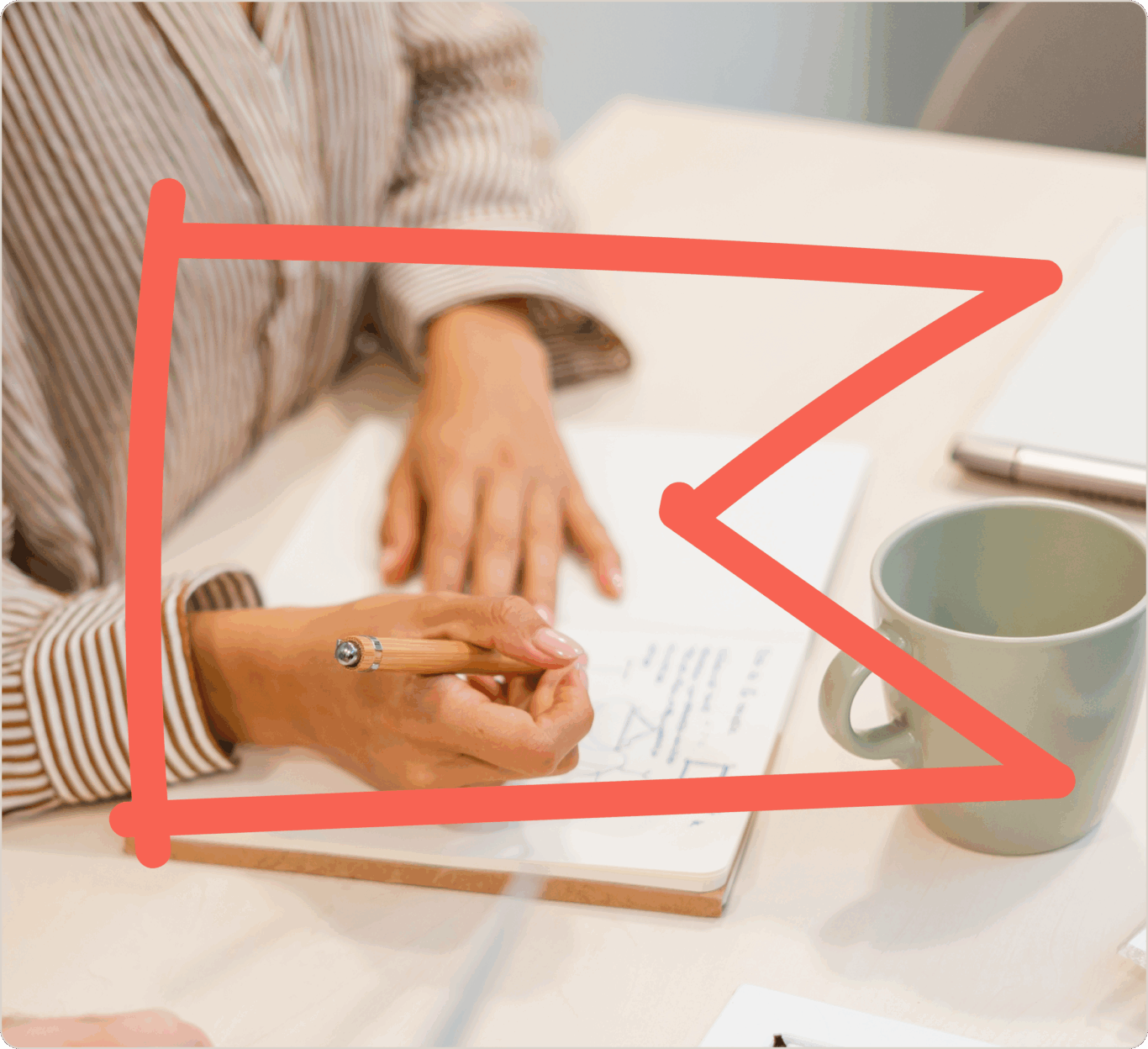 Person, die mit Bleistift in ein Notizbuch schreibt, daneben eine grüne Tasse auf einem Tisch. Ein roter Pfeil zeigt auf das Notizbuch.