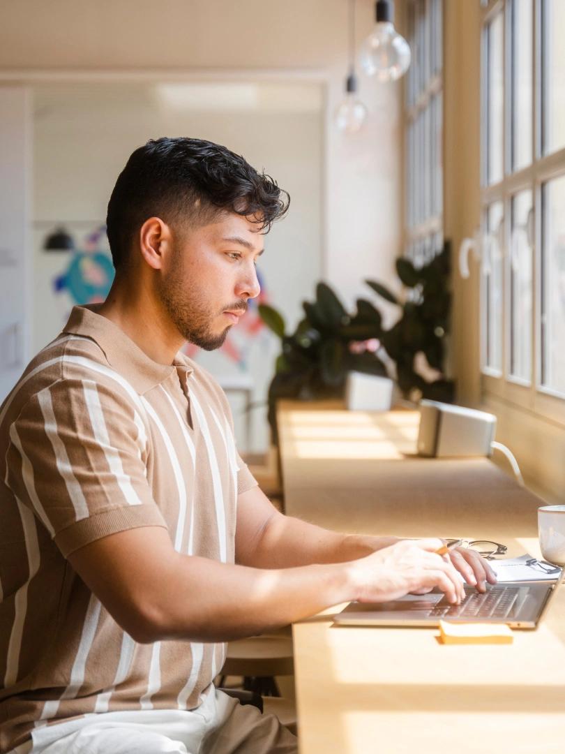 Man working on a laptop at a sunlit desk beside large windows.