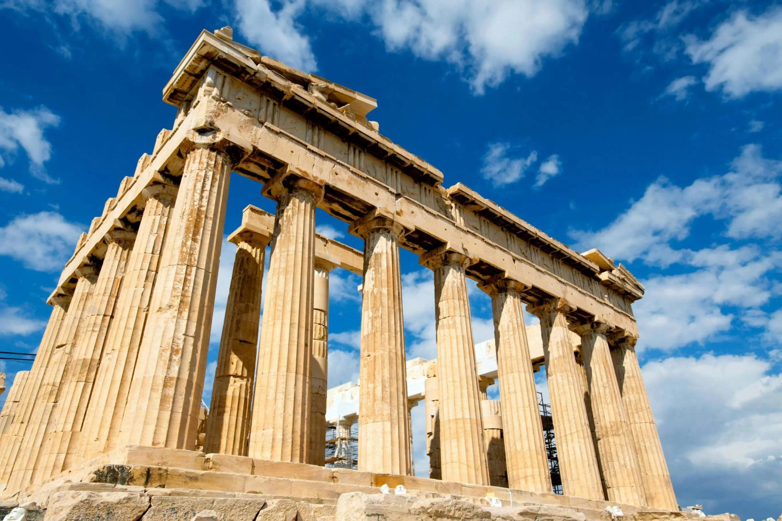 Vue du Parthénon à Athènes, un temple antique aux colonnes majestueuses en pierre dorée, se dressant sous un ciel bleu parsemé de nuages blancs.