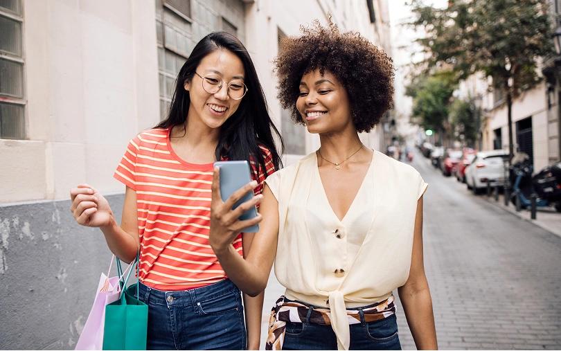 Two smiling women look at a smartphone while walking with shopping bags on a city street.