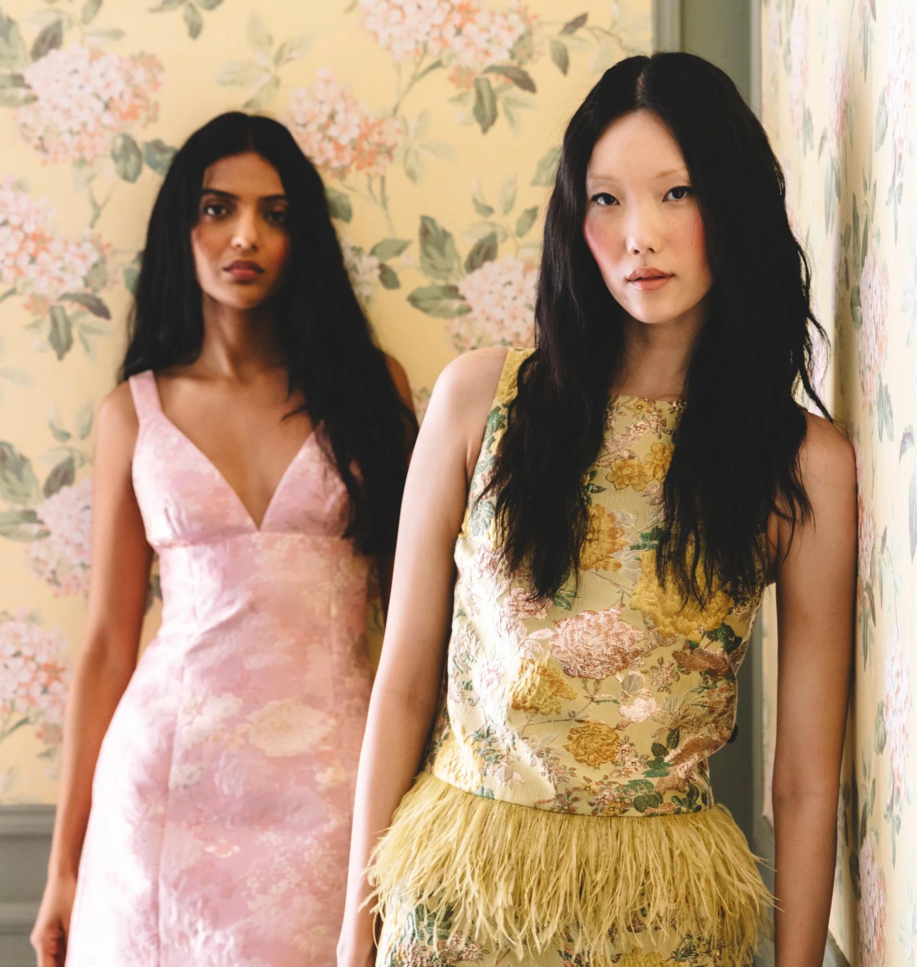 Two women model floral dresses against a coordinating flower-patterned wall.