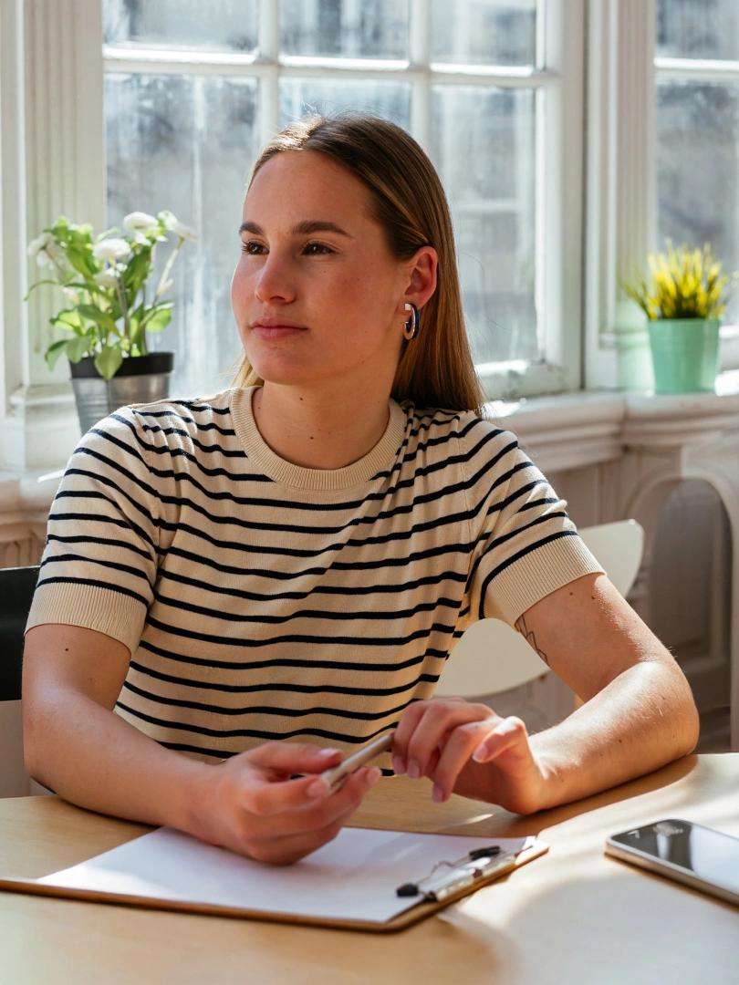 Woman holding a pen over a clipboard at a sunlit table, with a smartphone nearby.