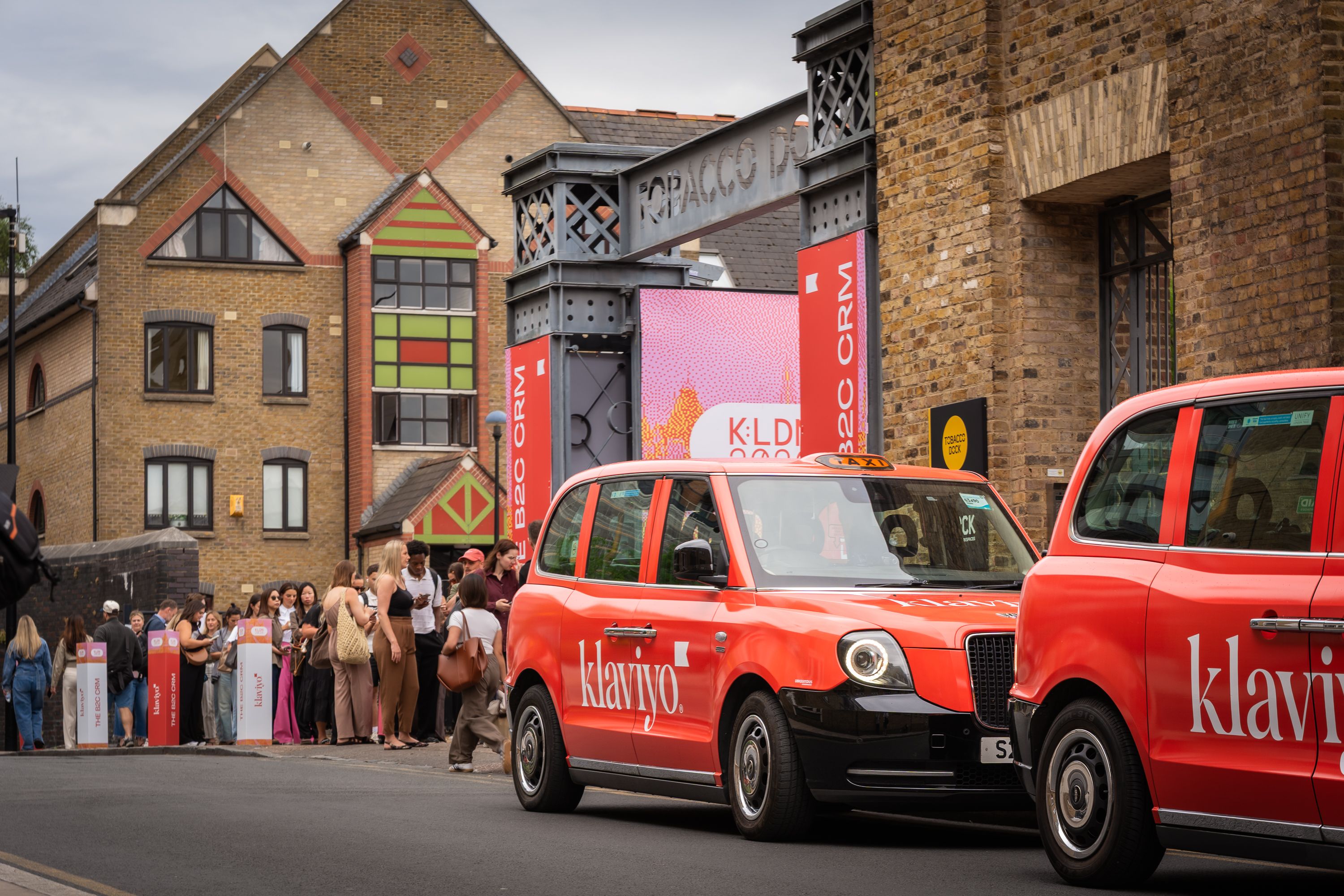 Two red "Klaviyo" taxis drive past a crowd waiting outside a building with event banners.