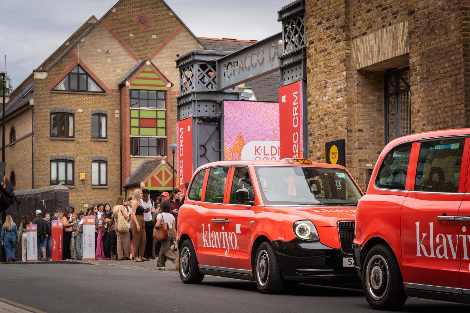 Two red 'Klaviyo' taxis drive past a crowd waiting outside a building with event banners.