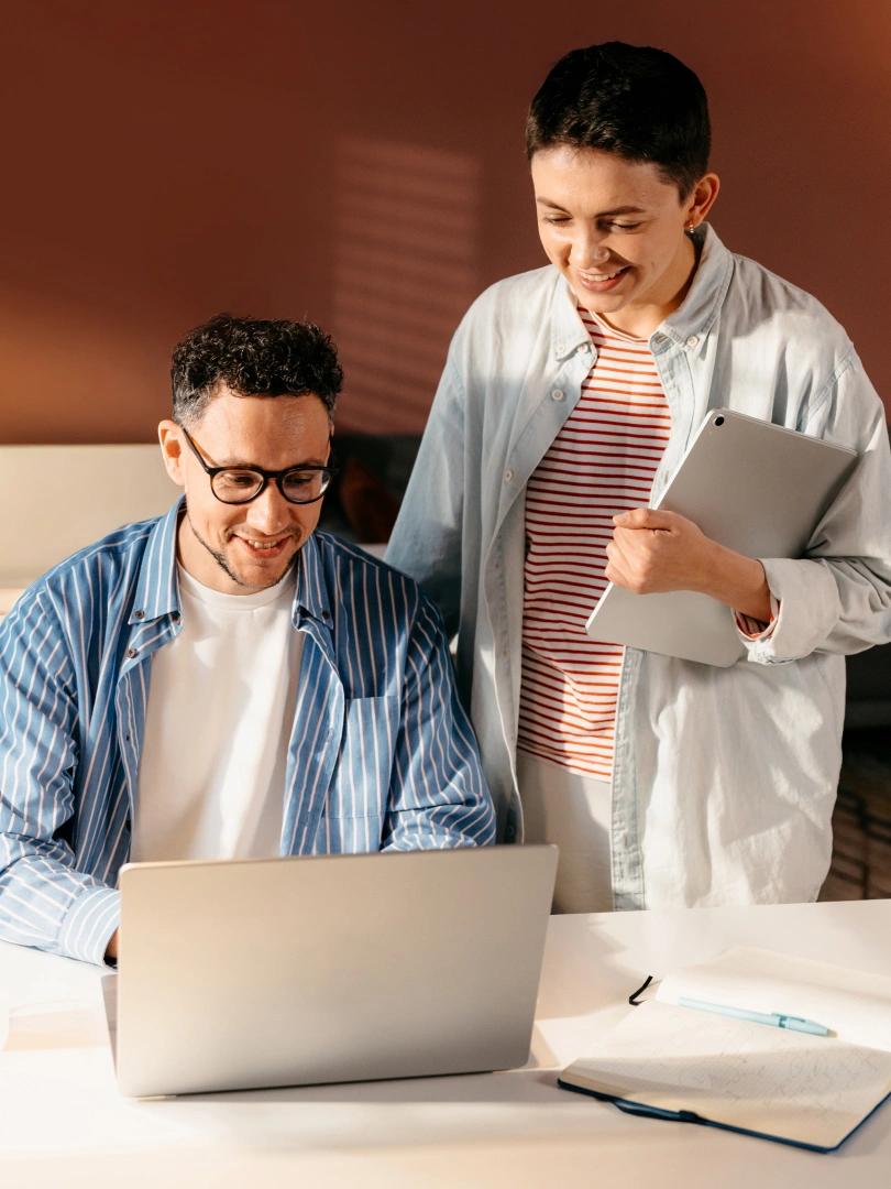Two smiling coworkers review a laptop together while one holds a tablet.