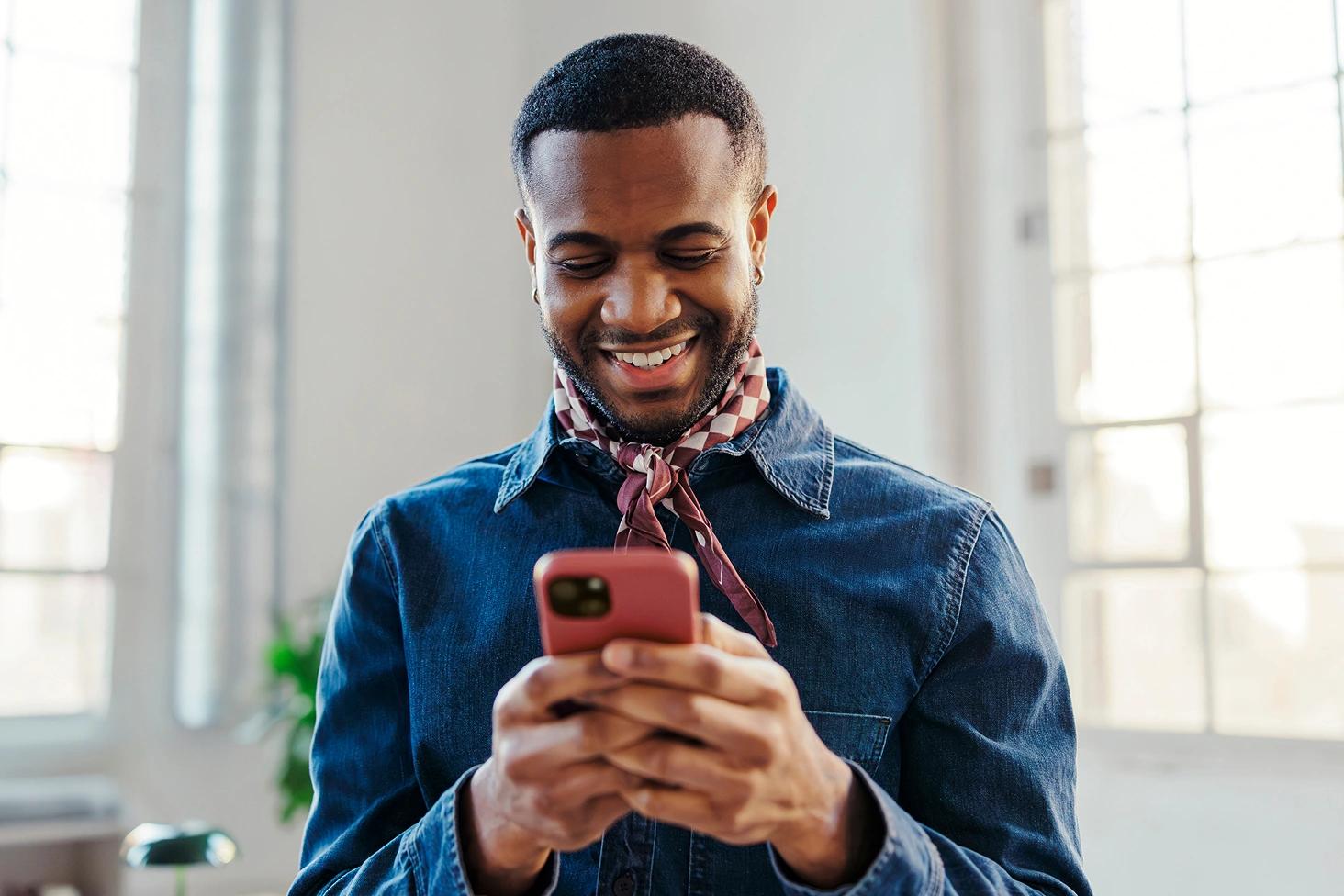 Smiling man in a denim shirt looks down at his smartphone in a bright room.