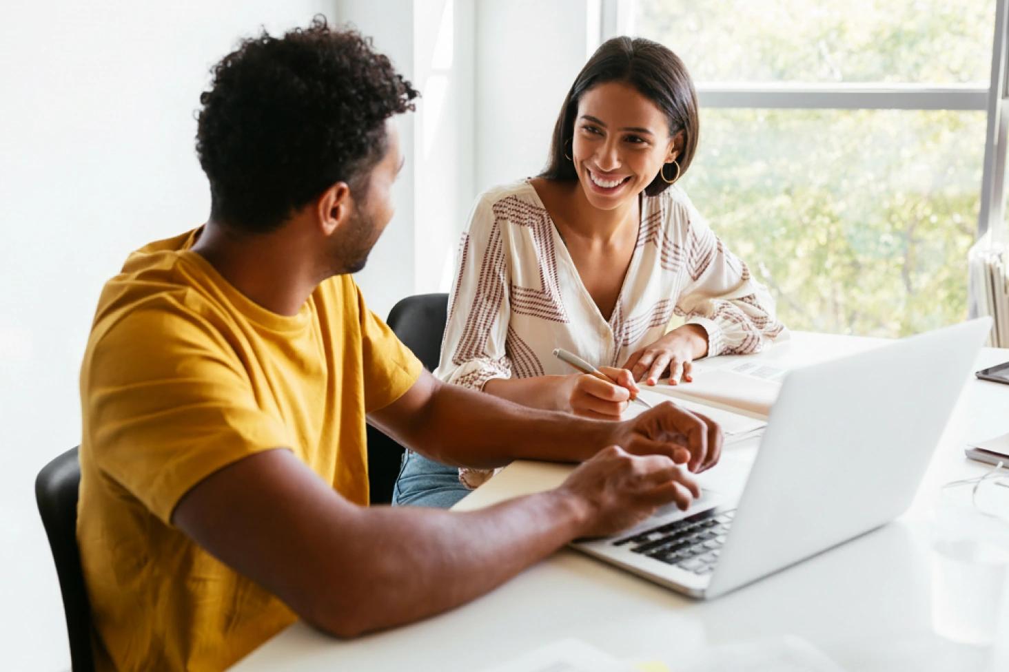 Two smiling coworkers collaborate over a laptop and notebook at a bright office table.