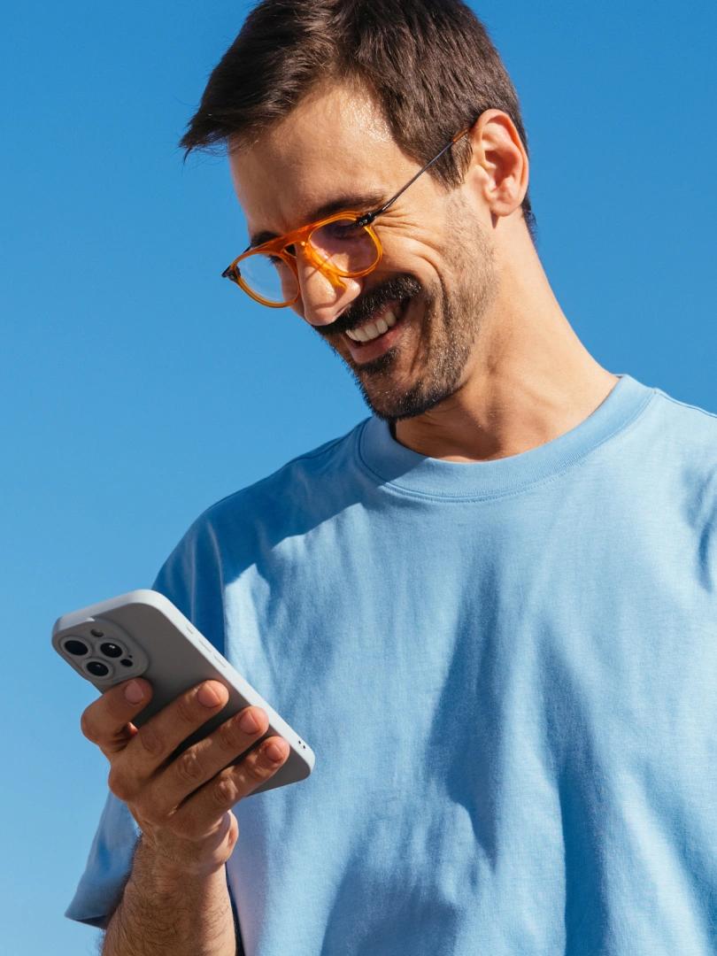 Smiling man in orange glasses and a blue shirt looks at his smartphone against a clear blue sky.