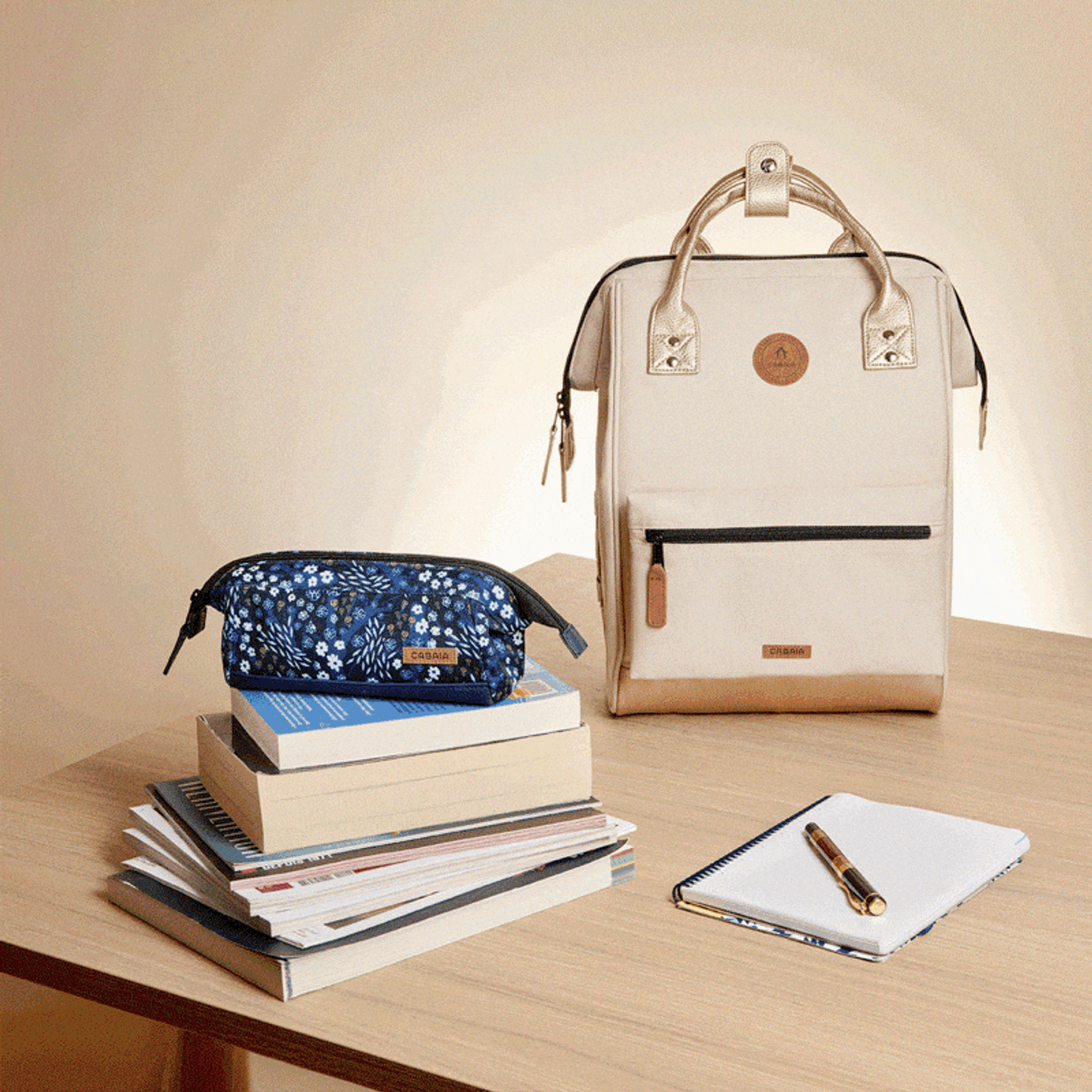 A cream and gold backpack, a blue floral pencil case, a stack of books, and a notebook with a pen on a wooden table.