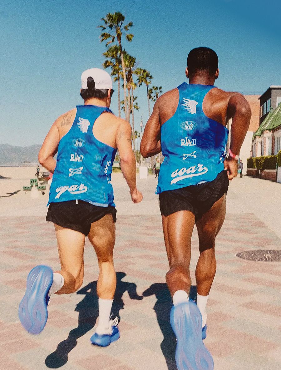 Two men in blue tank tops and black shorts run away from the camera on a sunny path with palm trees.
