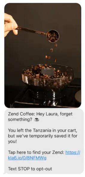 Image shows a smartphone text interface with one MMS inside. The product shot at the top shows a hand holding a small copper spoon, pouring coffee beans into a clear plastic receptacle for a grinder. Underneath a text message reads, “Zend Coffee: Hey Laura, forget something? You left Tanzania in your cart, but we’ve temporarily saved it for you! Tap here to find your Zend: https://kla6.io/0/8NFMWg. Text STOP to opt-out.”